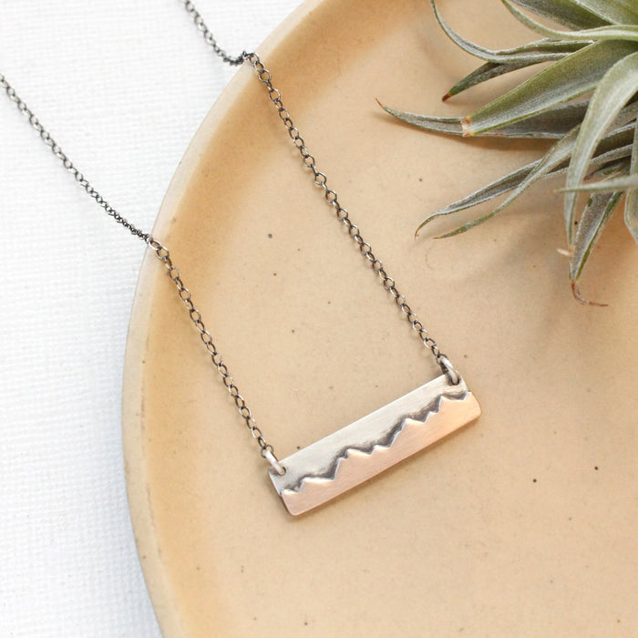 The oxidized silver layered mixed metal mountain bar necklace styled on a tan plate with an air plate