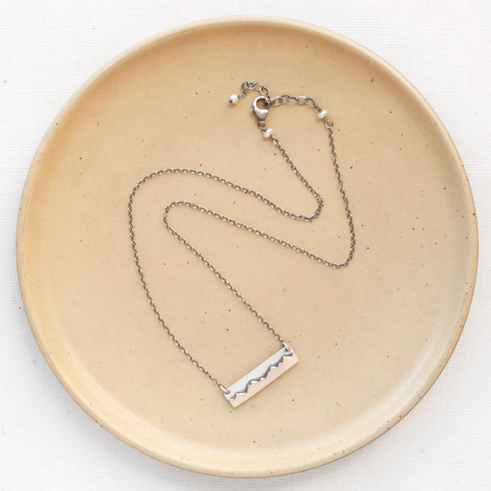 The layered mountain bar necklace styled on a tan plate to show the adjustable lobster clasp closure