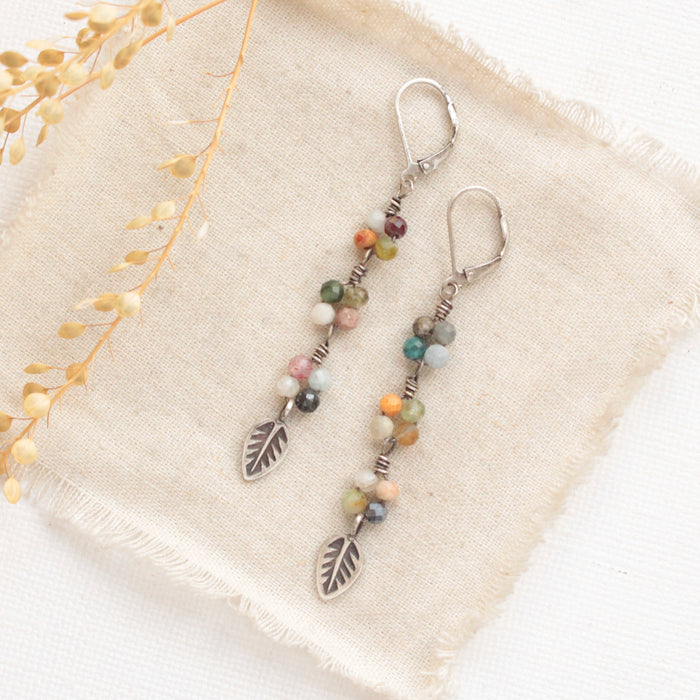 The long mixed gemstone leaf earrings styled on tan linen with dried grass