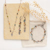 The kaleidscope leaf necklace, earrings, and bracelet styled with a tan rock, linen, and dried grass