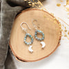 The mother of pearl and aquamarine hoop earrings styled on wood with gray velvet and dried grass
