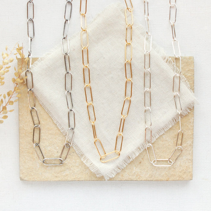 All three paperclip chain necklaces styled on a tan rock with linen and dried grass