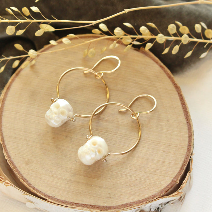 The carved pearl skull hoop earrings styled on wood with gray velvet and dried grass