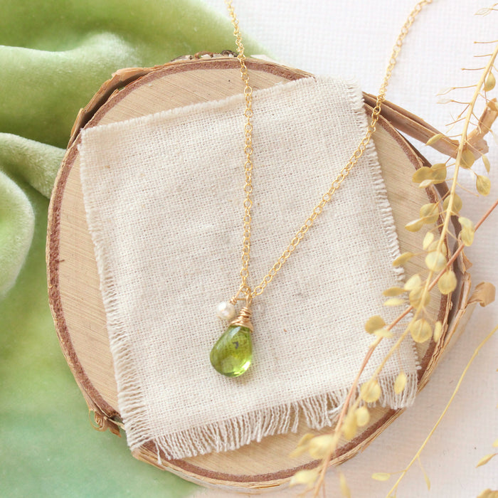 The peridot and pearl charm necklace styled on wood with green velvet, linen, and dried grass