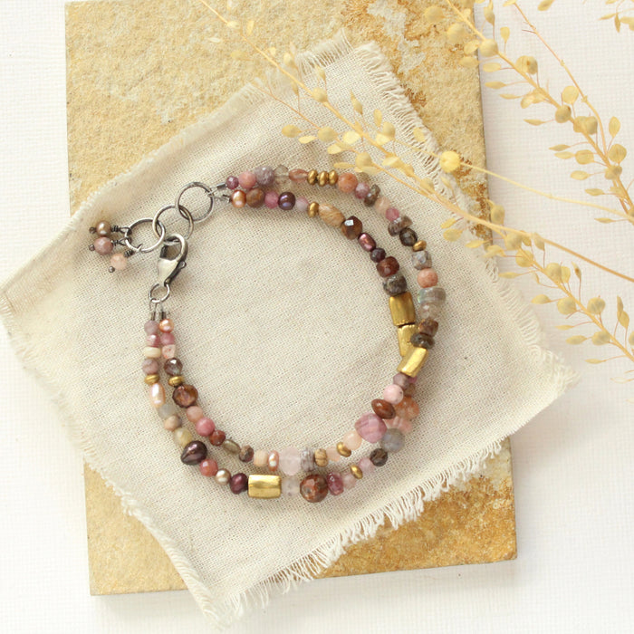 The pink and brass double strand bracelet styled on a tan rock with dried grass and linen