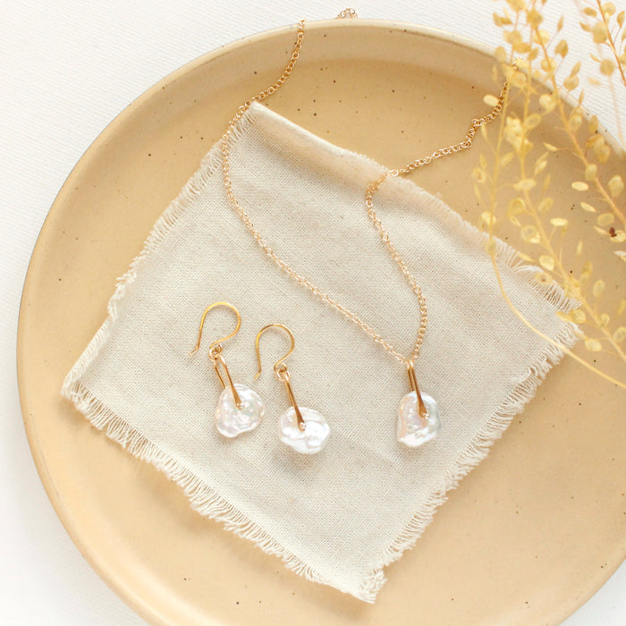 The pinned organic pearl earrings styled on a tan plate with the matching necklace, linen, and dried grass