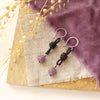 The ruby and leather earrings styled on a tan rock with linen, purple velvet, and dried grass