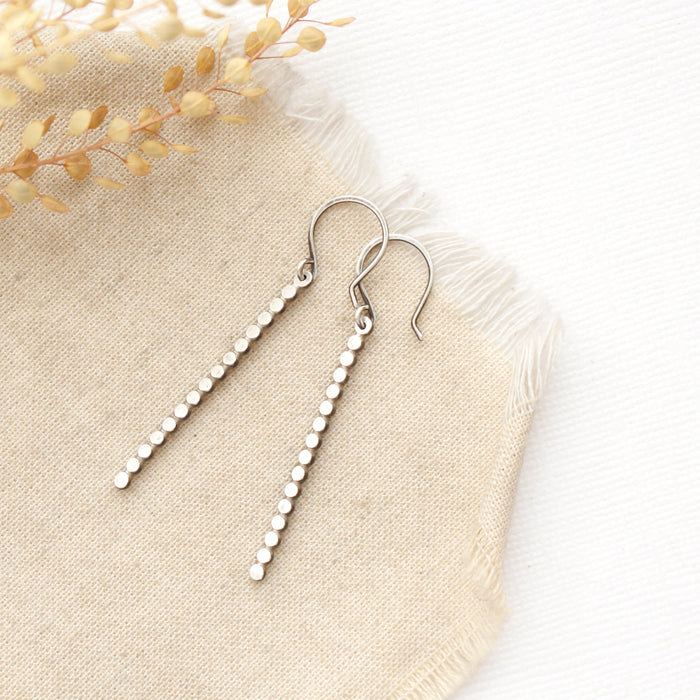 The silver beaded bar earrings styled on tan linen with dried grass