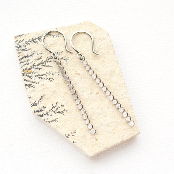 The silver beaded bar earrings styled on a tan rock