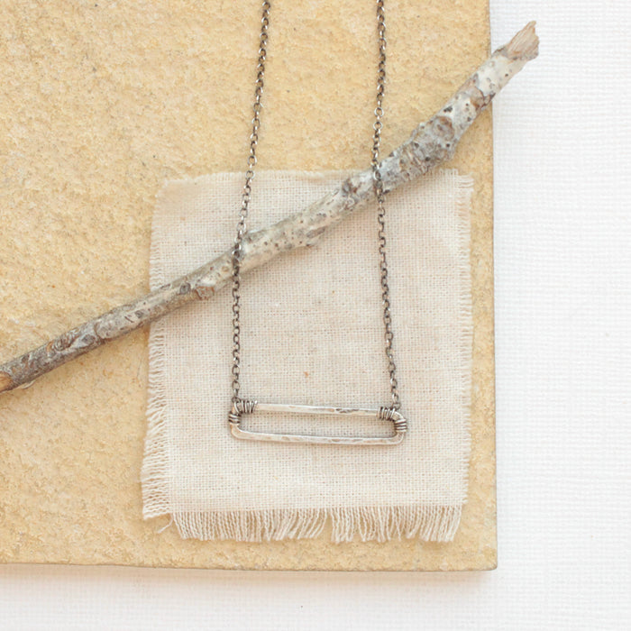 The silver open bar necklace styled on a tan rock with linen and a wooden stick