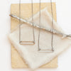 The silver open bar necklace and bronze open bar necklace styled together on a tan rock with linen and a wooden stick