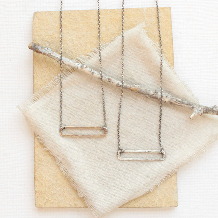 The silver open bar necklace and bronze open bar necklace styled together on a tan rock with linen and a wooden stick