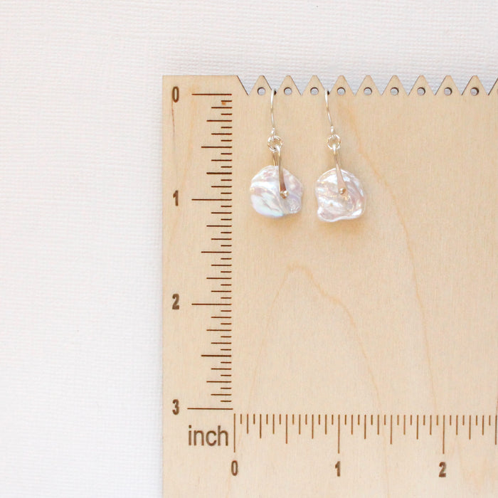 The pinned silver pearl earrings styled on a wooden ruler for size reference