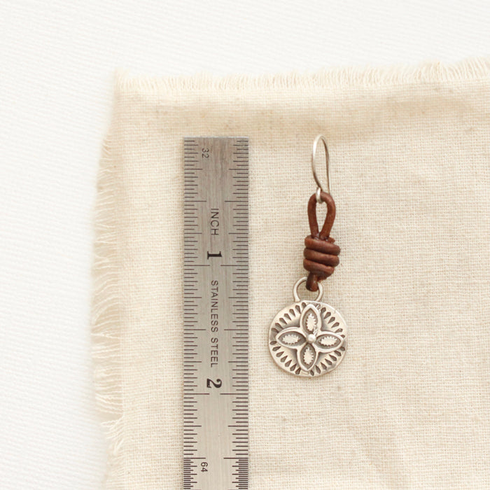 A silver wanderer coin leather earring next to a ruler for size reference
