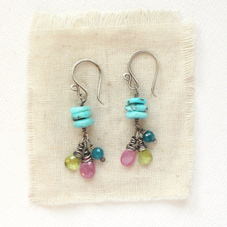 The colorful stacked turquoise, peridot, ruby, and apatite earrings styled on tan linen