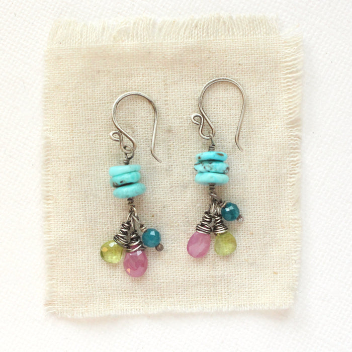 The colorful stacked turquoise, peridot, ruby, and apatite earrings styled on tan linen