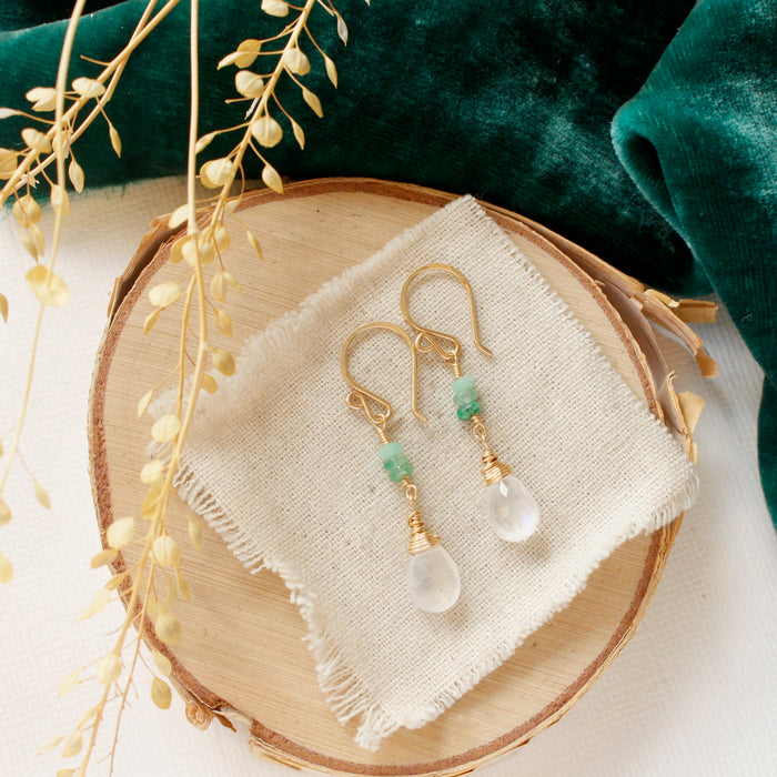 The stacked emerald and moonstone drop earrings styled on wood with green velvet and dried grass