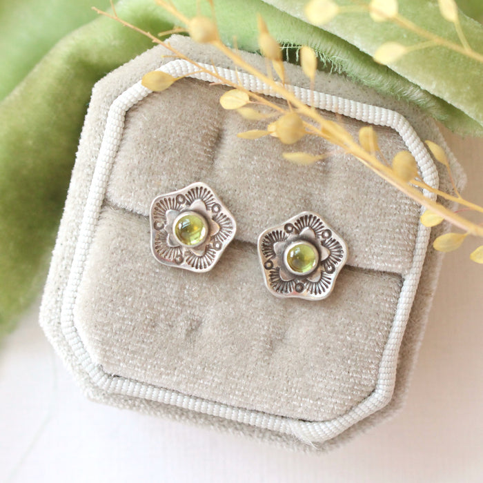 The stamped flower peridot post earrings styled on a white velvet box with green velvet and dried grass