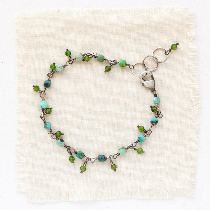 A bracelet made of turquoise cubes and dangling chrome diopside styled on tan linen.