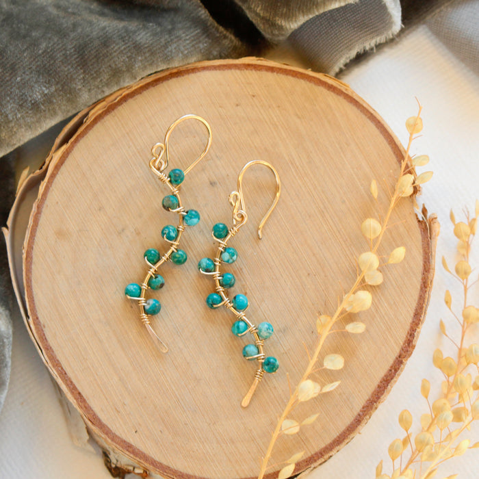 The turquoise wrapped gold vine earrings styled on wood with gray velvet and dried grass