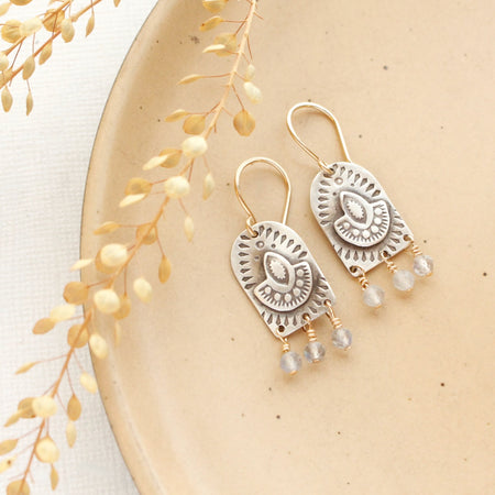 The wanderer arch mixed metal labradorite earrings styled on a tan plate with dried grass