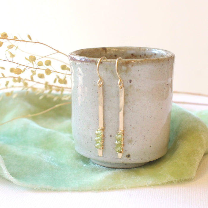 The peridot wrapped gold bar earrings styled on a ceramic cup with green velvet and dried grass