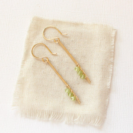 The peridot wrapped gold bar earrings styled on tan linen