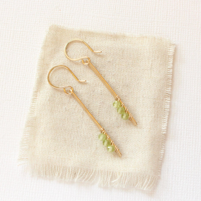 The peridot wrapped gold bar earrings styled on tan linen