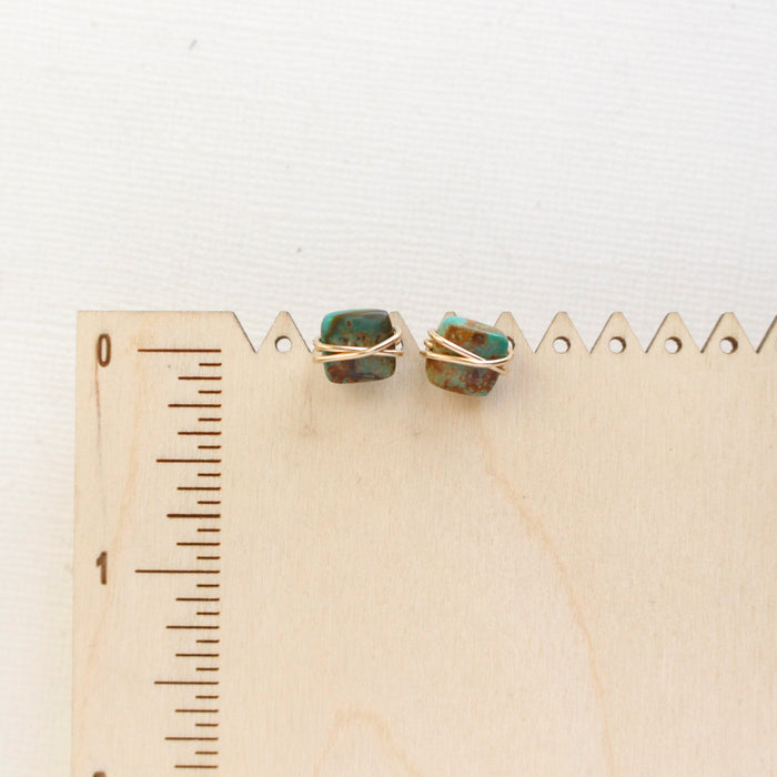 The gold wrapped turquoise square post earrings styled on a wooden ruler to show size reference