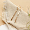 The yellow gold beaded bar earrings styled on tan linen with an air plant