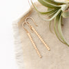 The yellow gold beaded bar earrings styled on tan linen with an air plant