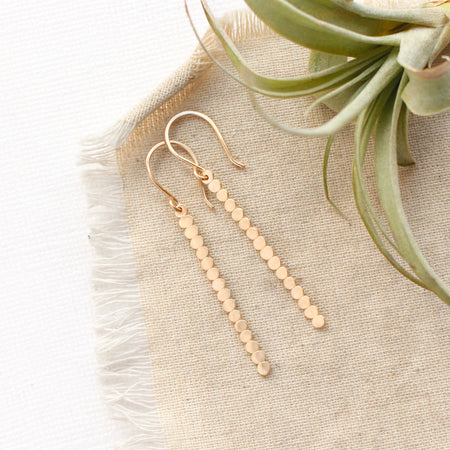 The yellow gold beaded bar earrings styled on tan linen with an air plant