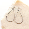 Forged oxidized silver teardrop hoop earrings styled on tan linen.
