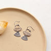 Stamped bronze and silver Asmi mixed duo earrings styled on a tan plate with a rock