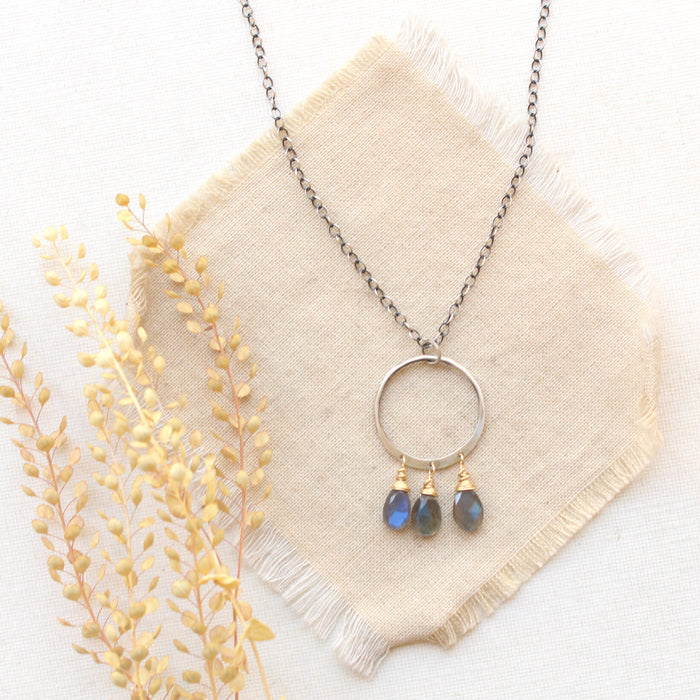 The forged labradorite trio hoop mixed metal long necklace is styled on tan linen with dried grass