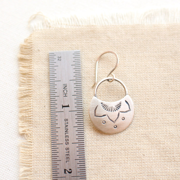 A stamped silver lotus half moon earring next to a ruler for size reference