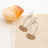 The stamped bronze and silver long loop Asmi earrings styled on tan linen with a crystal