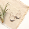 Stamped silver Asmi thin blade hoop earrings styled on tan linen with an airplant