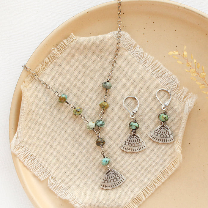 The asmi triangle turquoise lariat necklace styled in a tan plate with the matching earrings, linen, and dried grass
