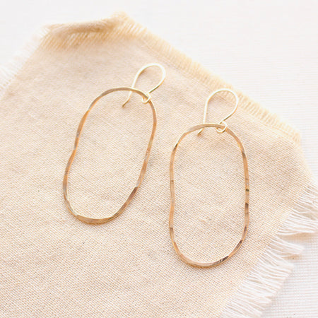 Gold organic shaped oval hoops with a hammered texture styled on tan linen.