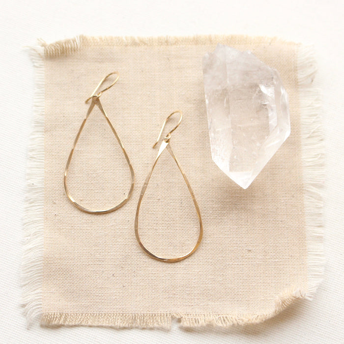 The yellow gold fill forged teardrop hoop earrings styled on tan linen with a crystal
