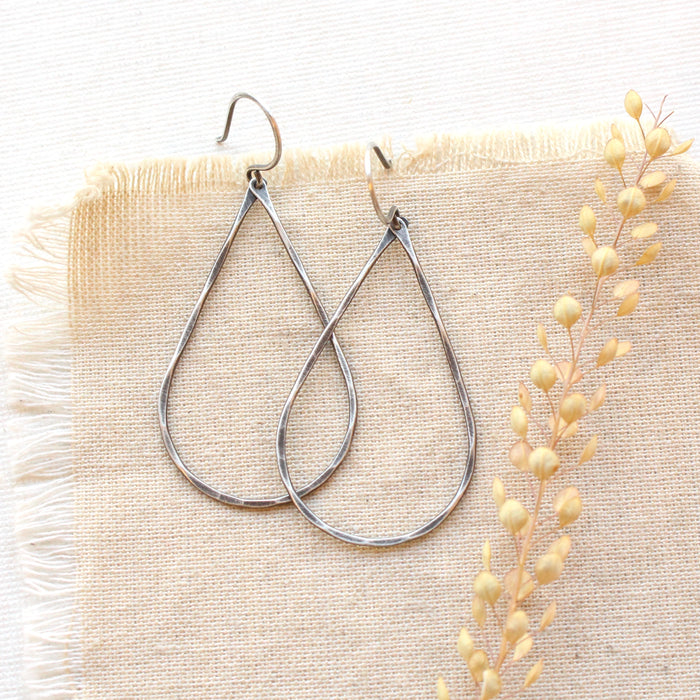 The oxidized silver forged teardrop hoop earrings styled on tan linen with dried grass