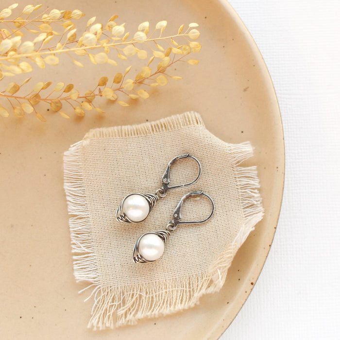 The oxidized silver perfect pearl earrings styled on a tan plate with linen and dried grass