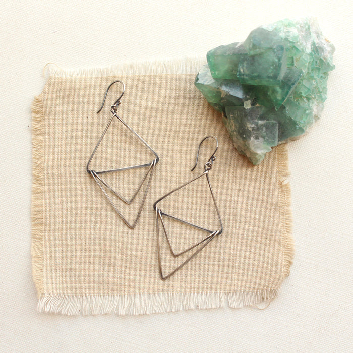 The oxidized silver triple triangle hoop earrings styled on tan linen with a green crystal