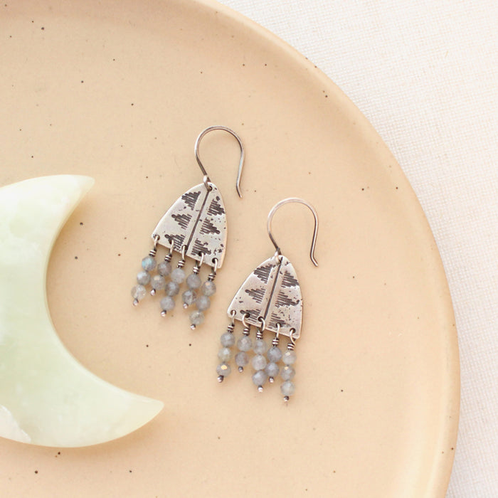 The pakal arch labradorite fringe earrings styled on a tan plate with a light green stone moon.