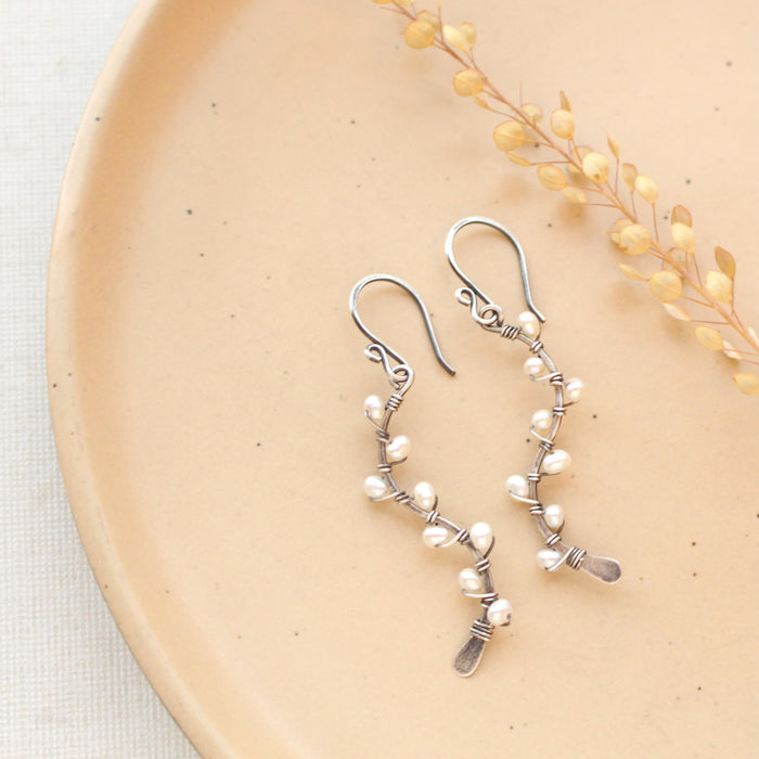 The pearl wrapped oxidized silver vine earrings styled on a tan plate with dried grass