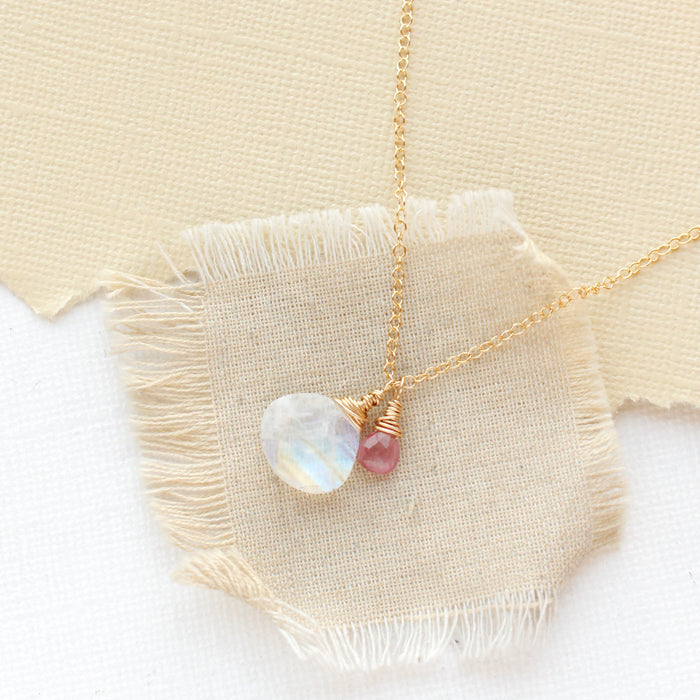 The pink sapphire and moonstone charm necklace styled on tan linen