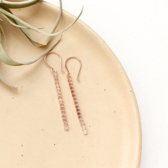 The rose gold beaded bar earrings styled on a tan plate with an air plant