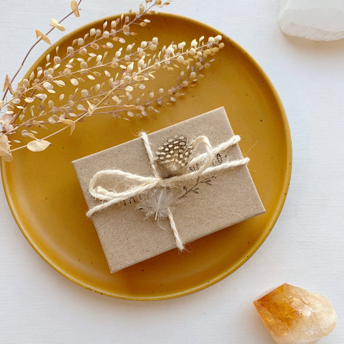 Sarah DeAngelo Jewelry box with signature gift wrap and feather styled on a yellow plate with dried grass and a crystal rock