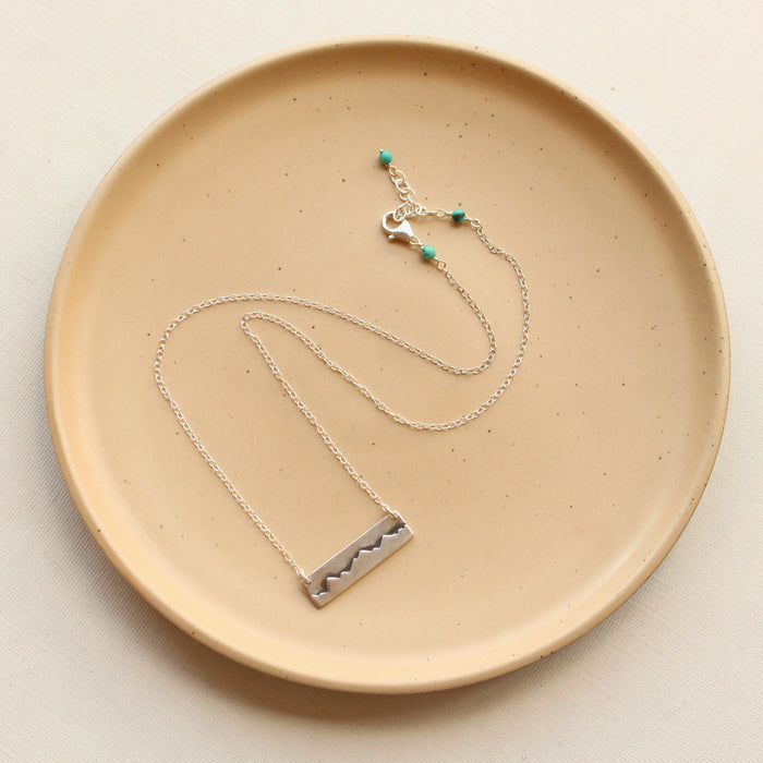 The silver layered mountain bar necklace styled on a tan plate to show the adjustable lobster clasp closure